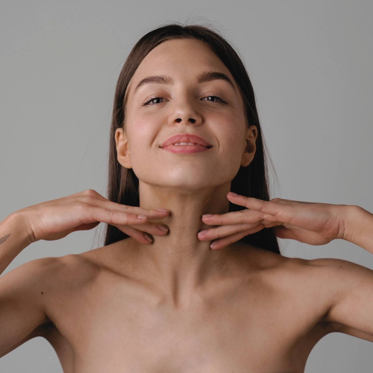 Noor naine teeb näojooga harjutust, hoides mõlemad käed lõua all, neutraalse taustaga.